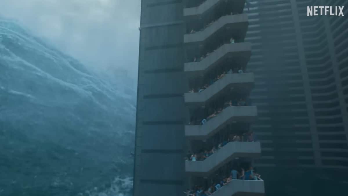 Cena do filme A Grande Inundação (Netflix 2025) mostrando onda gigante de água subindo ao lado de prédio com pessoas desesperadas nas escadas, representando o apocalipse por dilúvio.