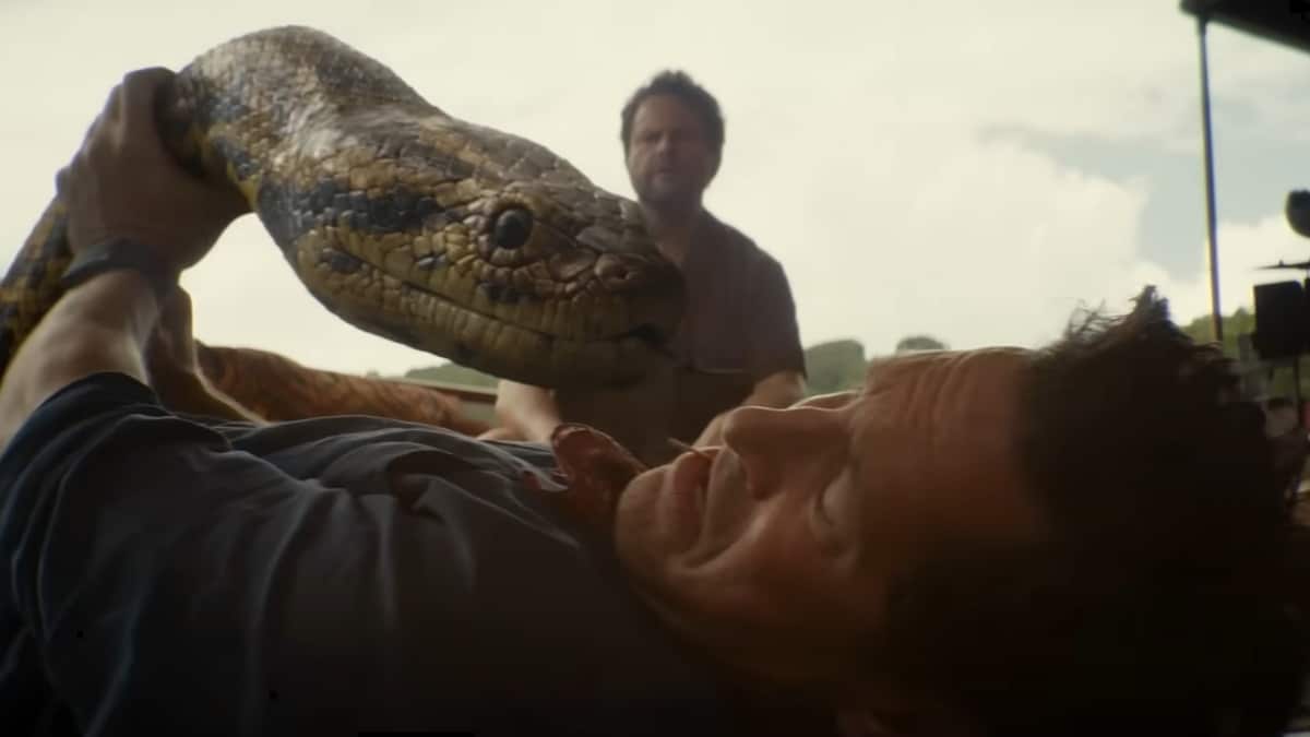 Cena do novo filme Anaconda mostra uma anaconda gigante se aproximando de um homem caído enquanto outro personagem observa ao fundo